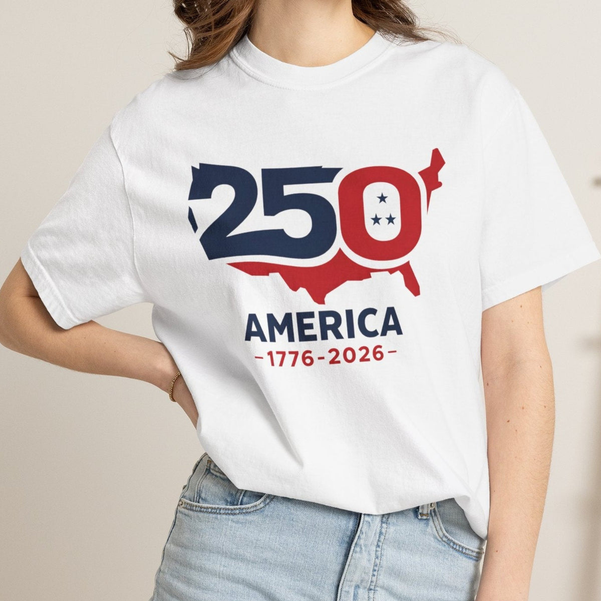 A woman wearing a white graphic t-shirt with a red and navy blue '250th Anniversary of America' design, paired with light-wash denim shorts, standing in a cozy indoor setting.