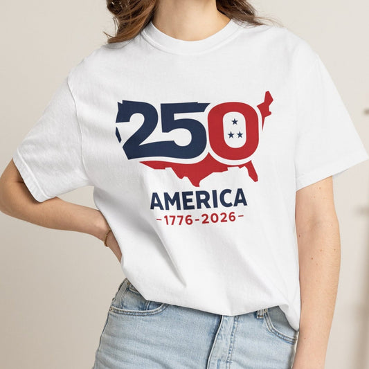 A woman wearing a white graphic t-shirt with a red and navy blue '250th Anniversary of America' design, paired with light-wash denim shorts, standing in a cozy indoor setting.
