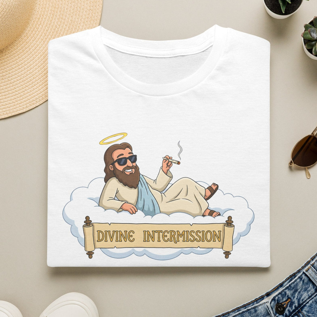 A white short-sleeve graphic T-shirt laid flat on a light beige surface, surrounded by a straw hat, white sneakers, blue jeans, small potted succulents, and sunglasses. The shirt features a cartoon illustration of a bearded man reclining on a cloud with a halo and a banner reading “DIVINE INTERMISSION.