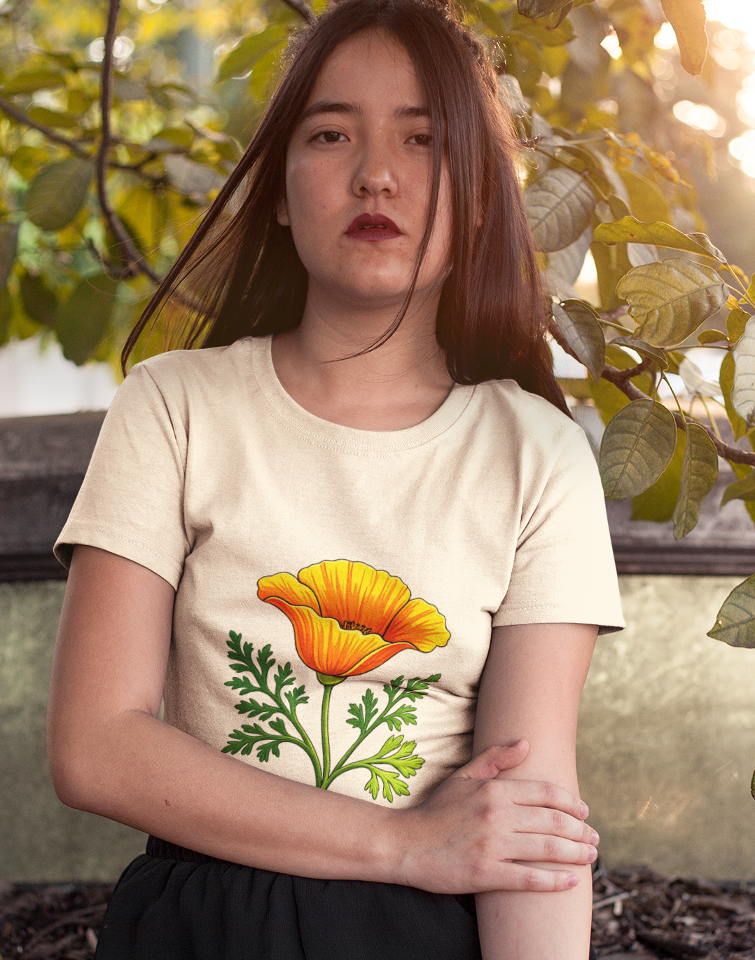 A young woman with long brown hair sits outdoors on a stone ledge, wearing a cream-colored t-shirt with a yellow and green poppy flower graphic on the front, paired with black sweatpants. Sunlight filters through the green leaves behind her.