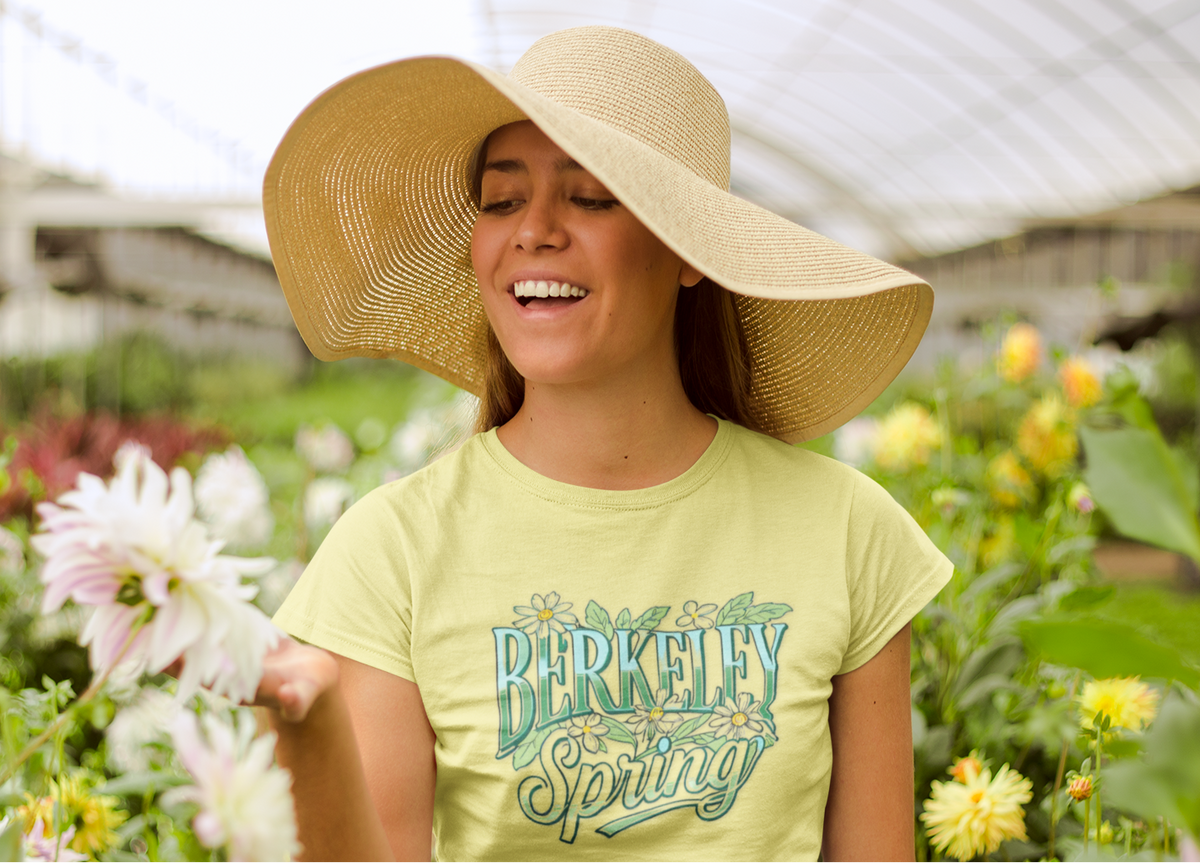 A woman wearing a large straw sun hat and a light yellow-green graphic T-shirt that reads 'Berkeley Spring' in teal, standing in a flower greenhouse surrounded by blooming dahlias.