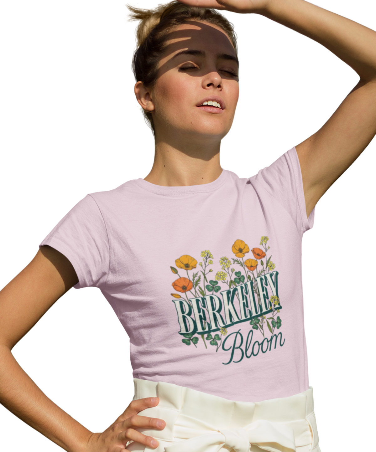 A woman wearing a light pink graphic t-shirt with 'Berkley Bloom' and a floral design, paired with cream-colored pants, against a solid white background.