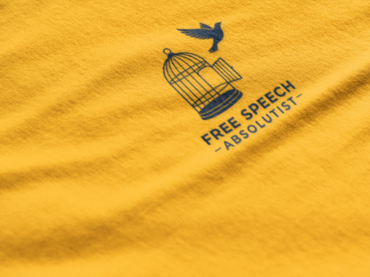 A close-up of a yellow fabric surface with a dark navy printed graphic and text reading “FREE SPEECH - ABSOLUTIST” beneath a birdcage and bird illustration.