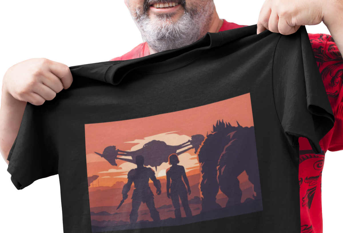 A smiling man with a gray beard holds up a black graphic t-shirt against a plain white background. The t-shirt features a rectangular print of silhouetted figures and creatures against an orange sunset sky.