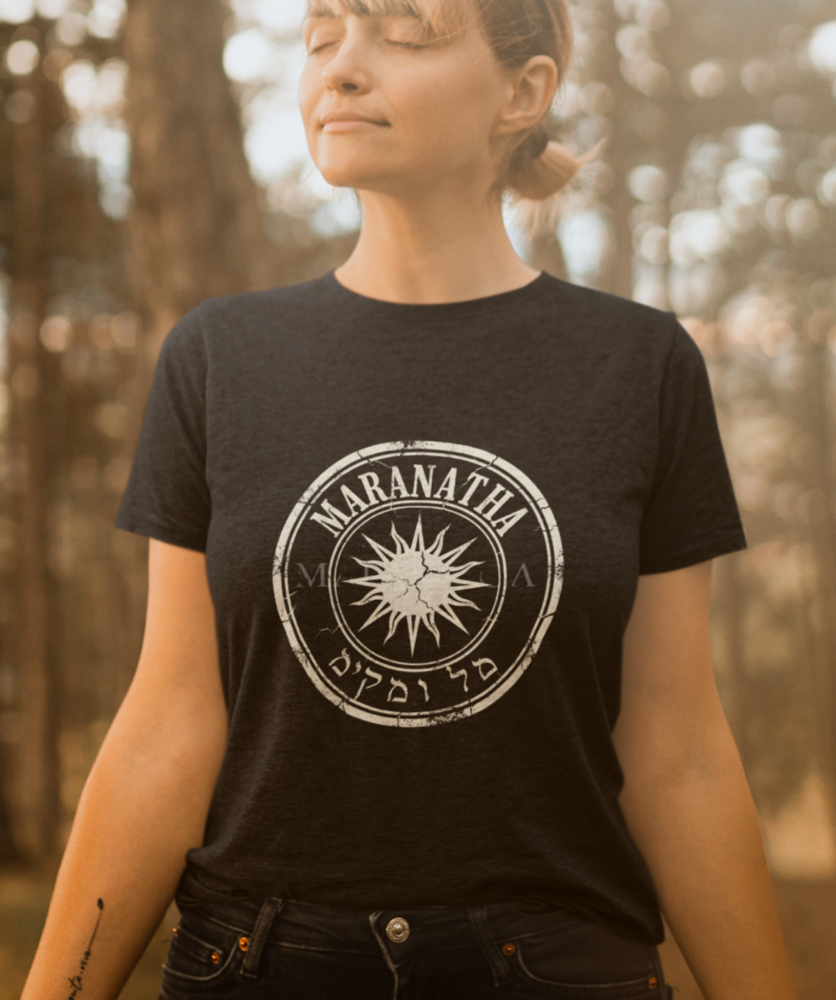 A woman stands in a sunlit forest with her eyes closed, wearing a black graphic T-shirt with a white circular sunburst design and the word 'MARANATHA' printed on the chest, paired with dark jeans.