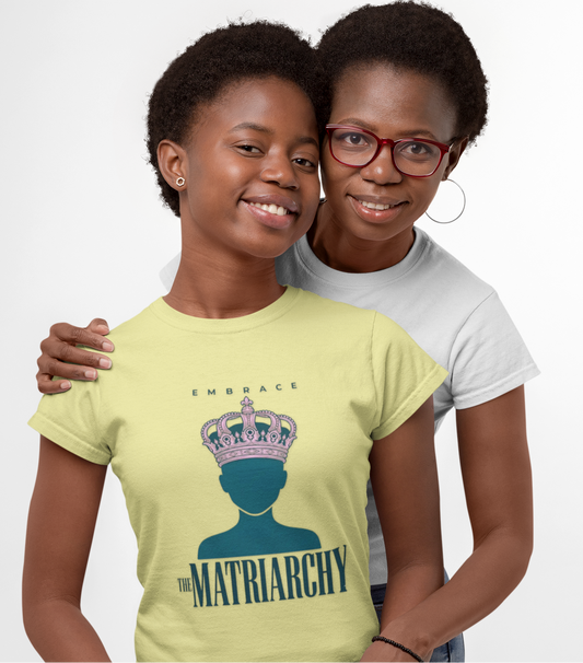 Two women posing together against a light gray studio background. The woman in front wears a pale yellow graphic T-shirt with a crown and the words 'EMBRACE THE MATRIARCHY' printed on the front, paired with blue jeans.
