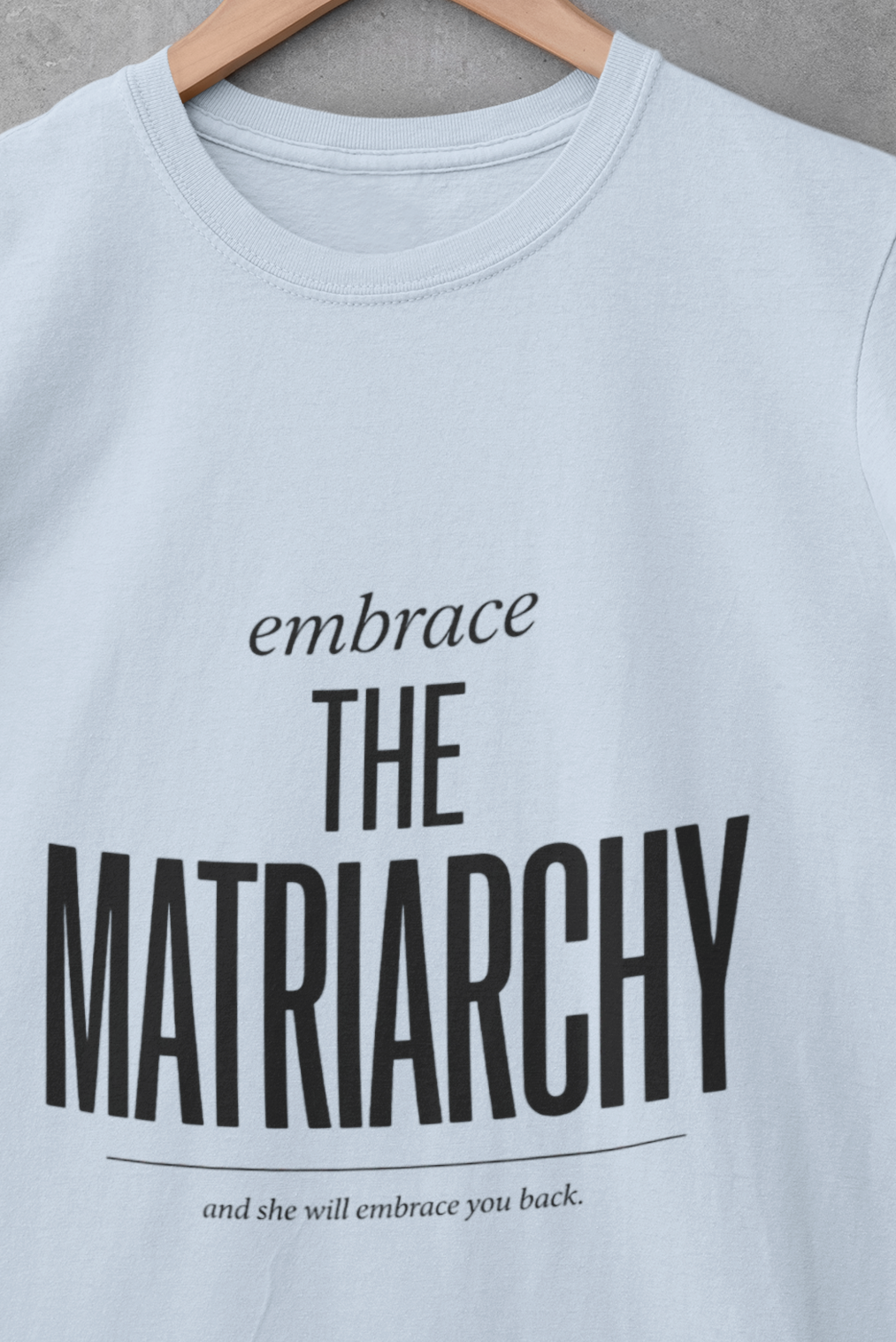 A light blue short-sleeve t-shirt hanging on a wooden hanger against a textured gray wall. The shirt features a black printed graphic that reads 'embrace THE Matriarchy' with a smaller line of text below reading 'and she will embrace you back.