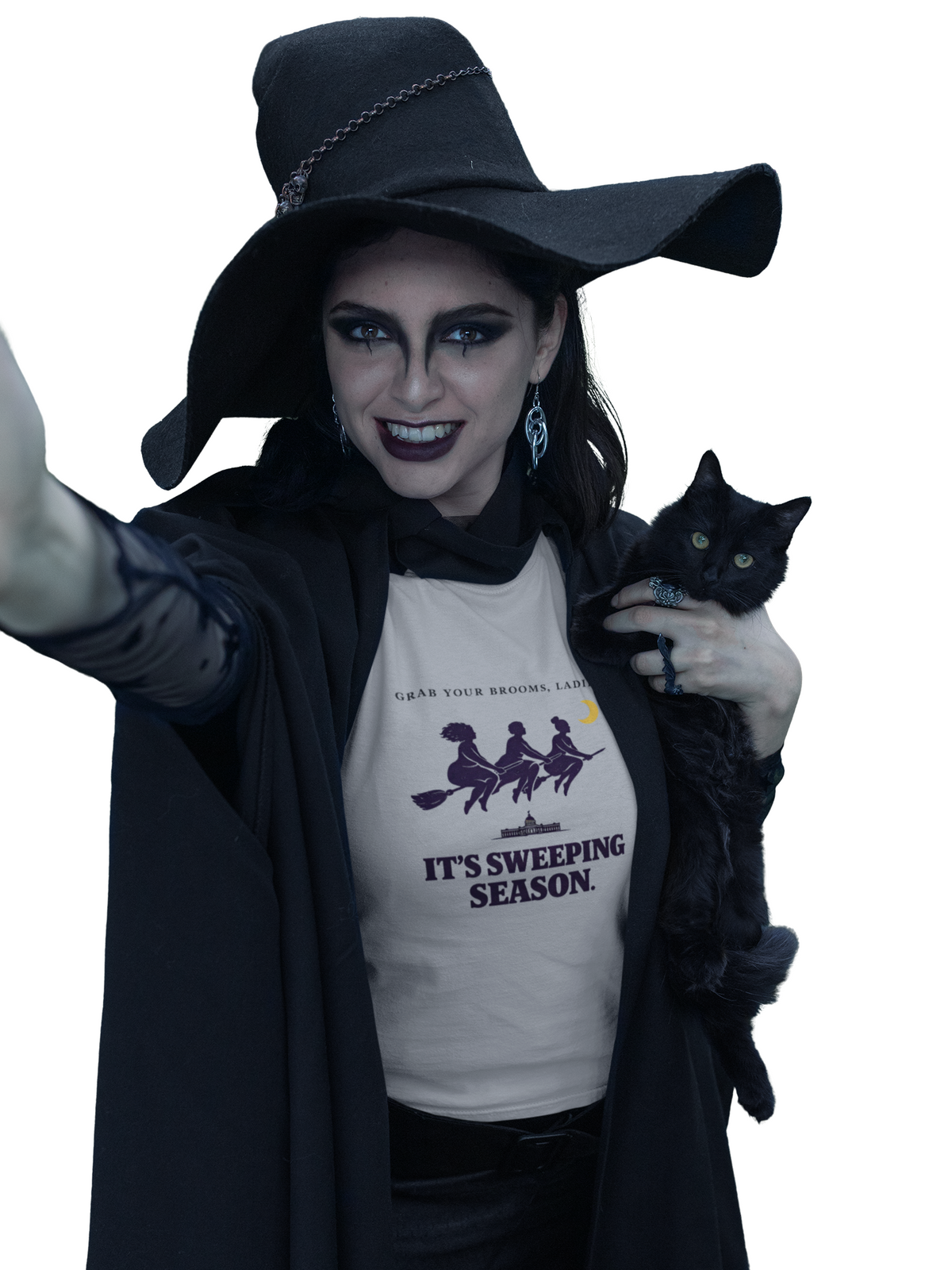 A woman in a witch costume with a large black hat, black cloak, and face paint holds a black cat. She is wearing a white graphic t-shirt that reads 'IT'S SWEEPING SEASON.