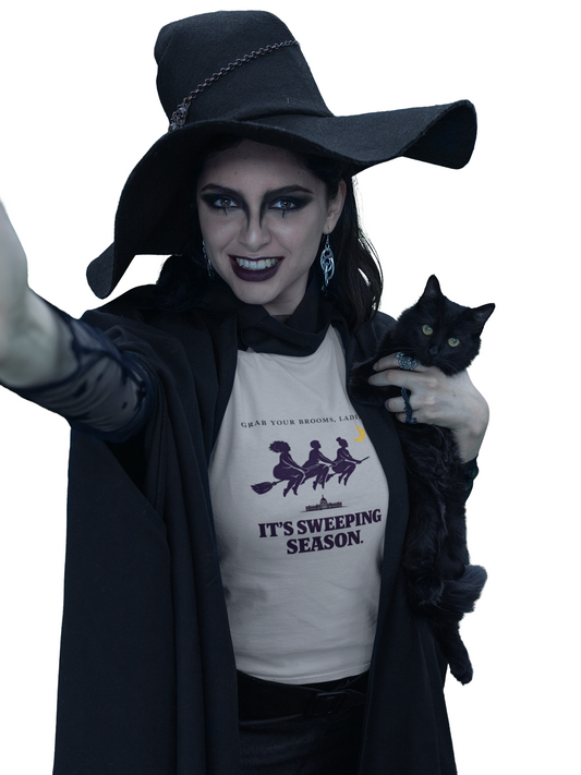 A woman in a witch costume with a large black hat, black cloak, and face paint holds a black cat. She is wearing a white graphic t-shirt that reads 'IT'S SWEEPING SEASON.