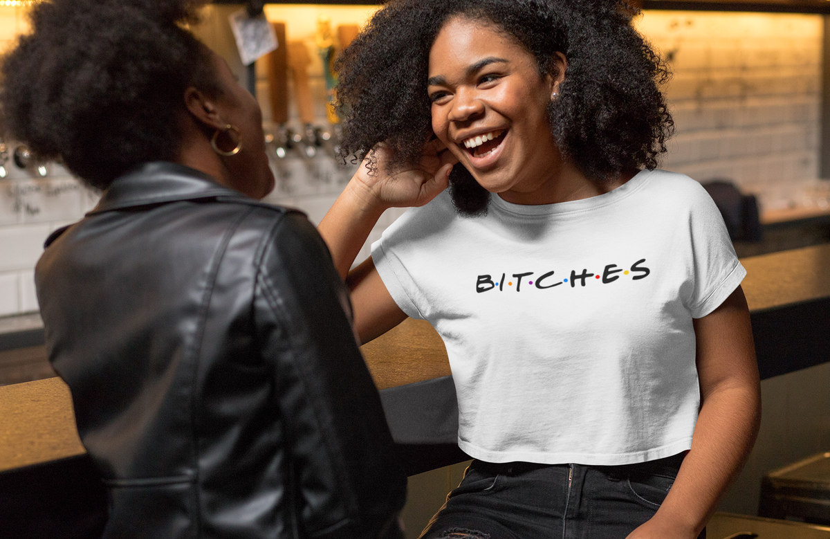 A woman in a white graphic t-shirt with the word 'BITCHES' printed in black across the chest, laughing while sitting in a bar or cafe setting next to another person.