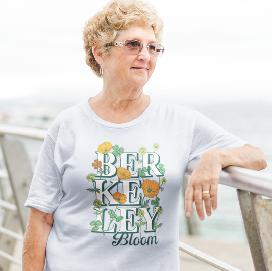 An older woman with short curly blonde hair and glasses is wearing a light blue crewneck t-shirt with a colorful graphic that reads 'BERKEYLEY Bloom' surrounded by flowers and leaves. She is leaning on a metal railing outdoors with a blurred background.