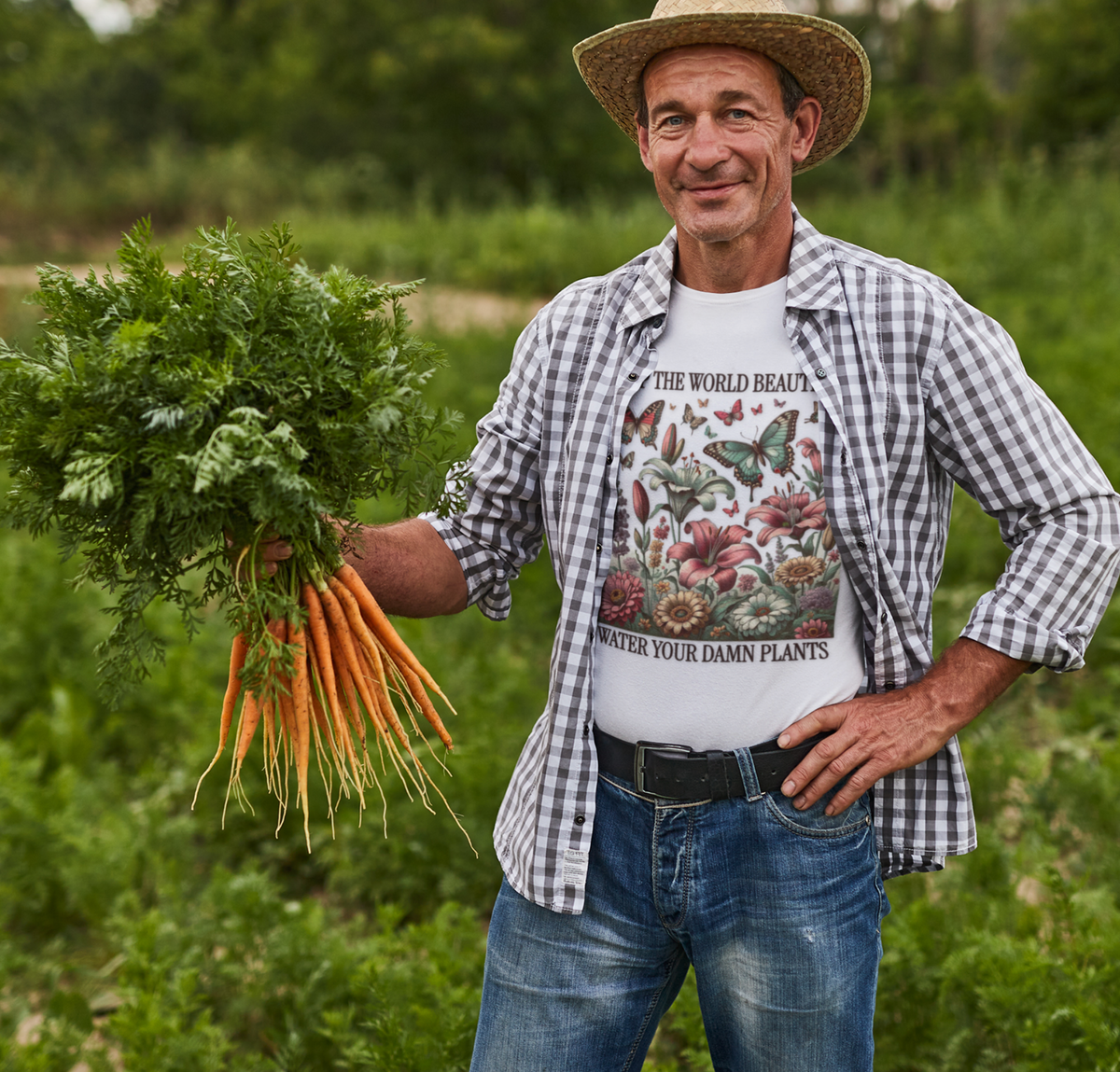 A man wearing a straw hat, a plaid button-up shirt, and a graphic t-shirt stands in a green field holding a large bunch of freshly harvested carrots with green tops.