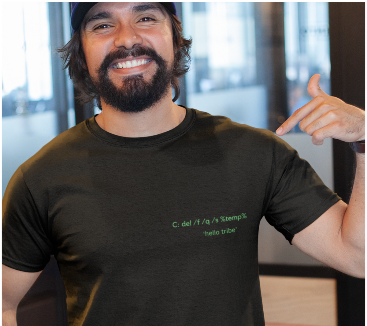 A smiling man wearing a dark charcoal gray t-shirt with green text on the chest that reads 'C. del H / a / r's 'stempli', 'hello tribe'.