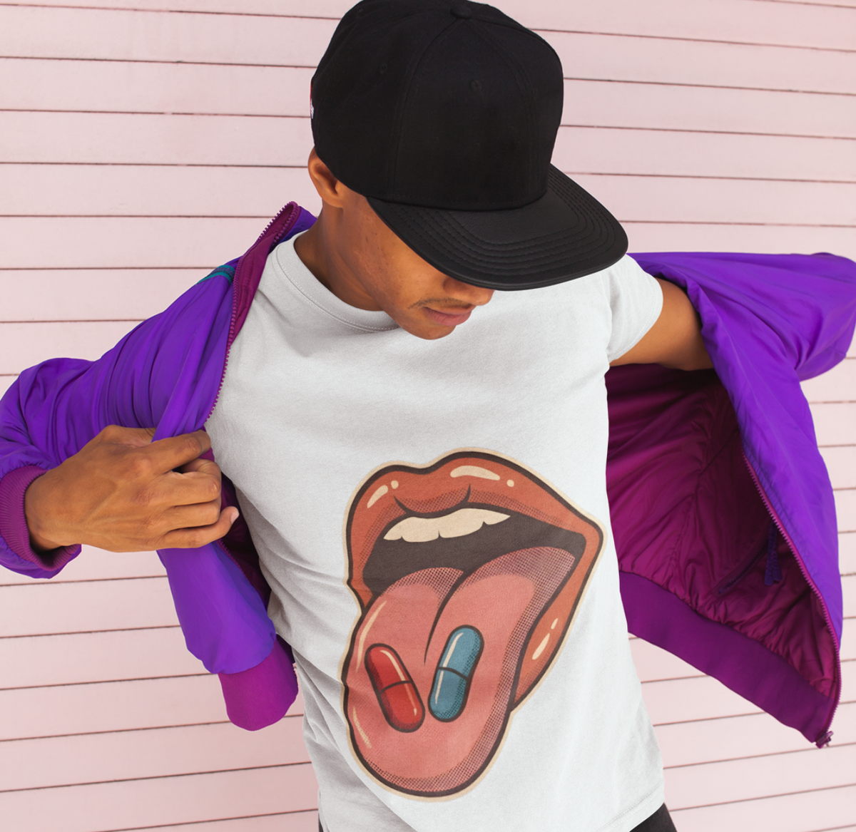 A person wearing a white graphic t-shirt with a tongue-and-dots design, a black baseball cap, and a purple puffer jacket draped off the shoulders, standing against a pink slatted wall.