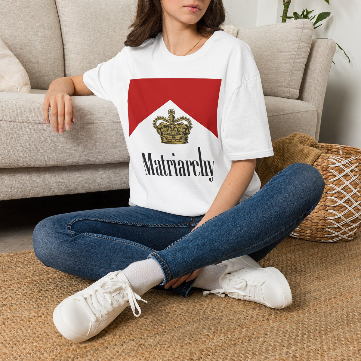 A woman sitting cross-legged on a woven rug in front of a beige sofa, wearing a white graphic T-shirt with a red and white flag design and the word 'Matriarchy' in black, paired with blue jeans and white sneakers.