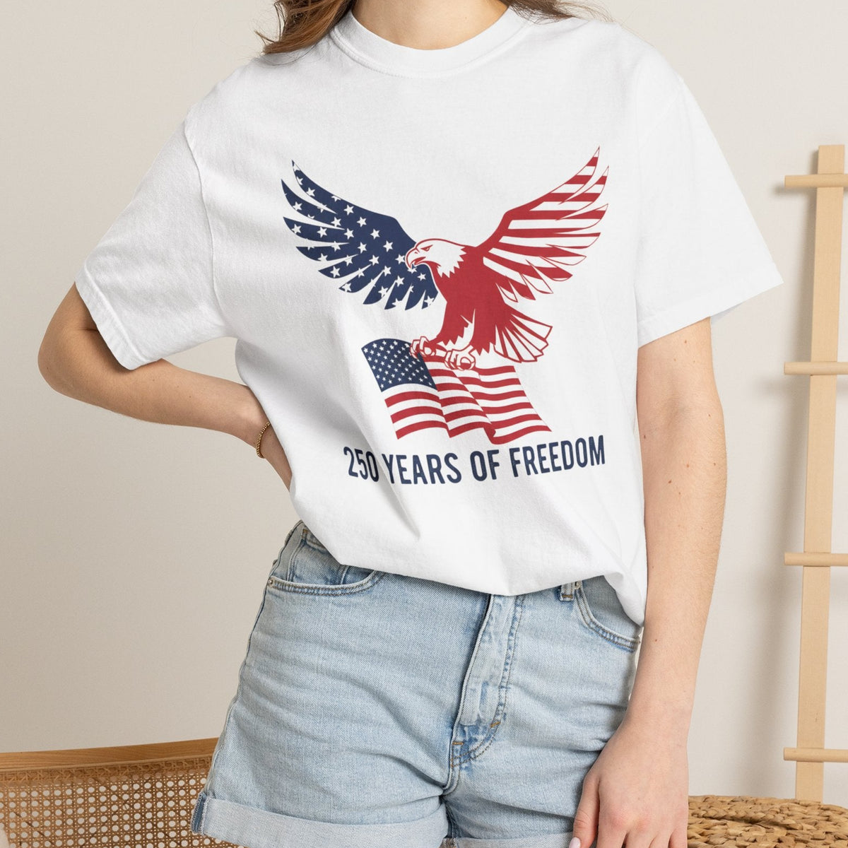 A woman wearing a white short-sleeve graphic T-shirt with a red, white, and blue eagle and American flag design, paired with light blue denim shorts, in a softly lit indoor setting.