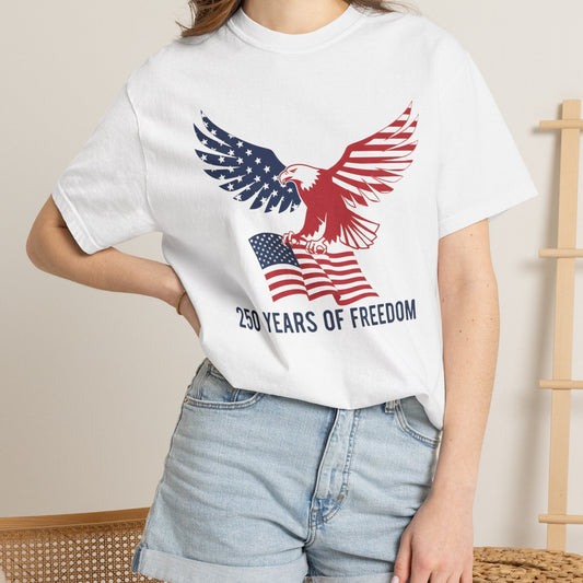 A woman wearing a white short-sleeve graphic T-shirt with a red, white, and blue eagle and American flag design, paired with light blue denim shorts, in a softly lit indoor setting.