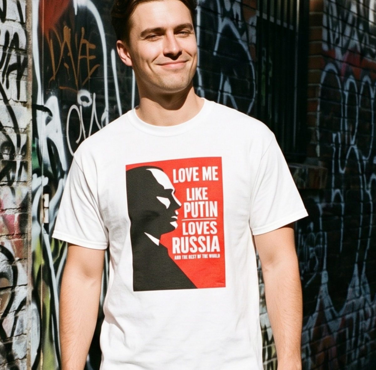 A man wearing a white graphic t-shirt with a red and black print that reads 'LOVE ME THE PUTIN LOVES RUSSIA' and black pants, standing in an outdoor alleyway with graffiti-covered brick walls.