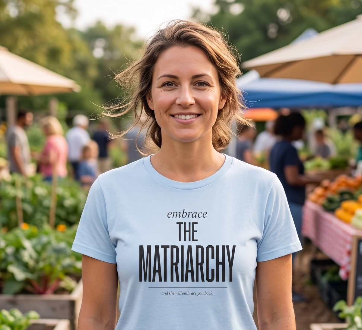 A woman stands in a farmers market wearing a light blue graphic t-shirt with the text 'embrace THE Matriarchy' printed on the front. The background shows blurred rows of garden beds and market stalls.