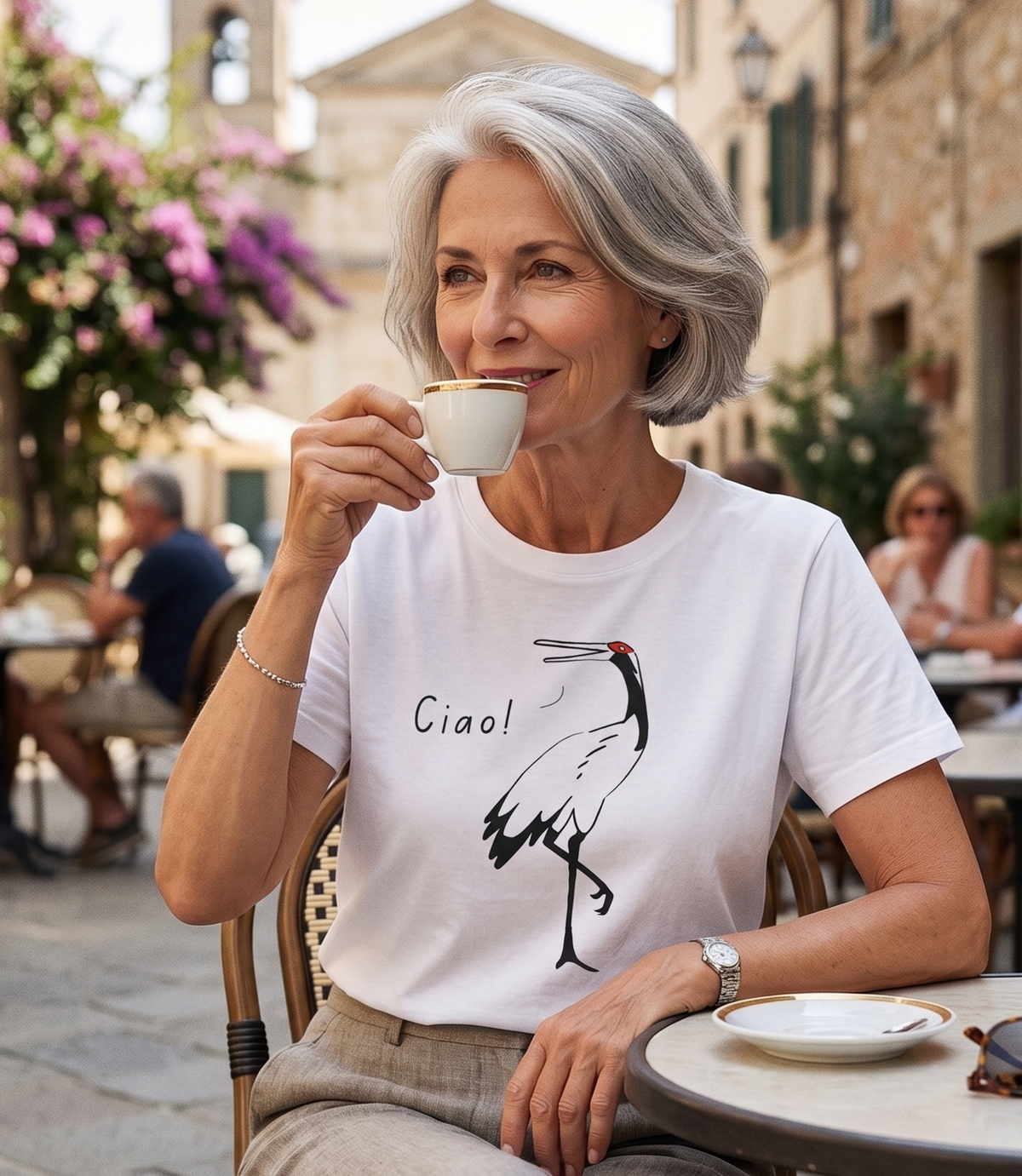 A woman with gray hair sits at an outdoor cafe table, holding a white cup. She is wearing a white graphic t-shirt with a black stork illustration and the text 'Ciao!' on the front, paired with beige trousers.